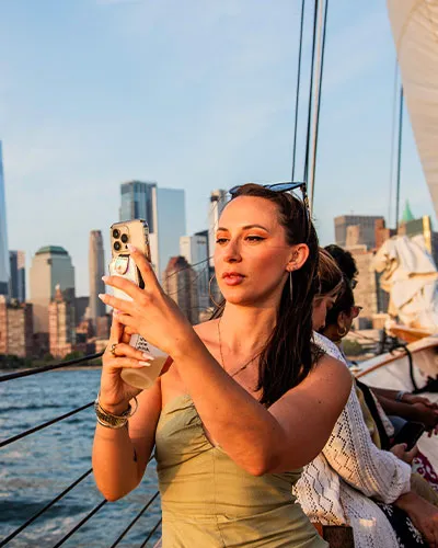 closeup of a woman holding a drink, with a phone in her hand looking out over the water on a sailboat