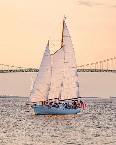 a classic harbor line sailboat cruising across the water with a giant arch bridge behind it