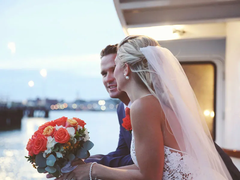 a man and a woman leaning out over a motor yacht, dressed for their wedding, while the woman is holding flowers